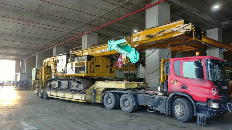 A red Hong Li Scania prime mover with a yellow crawler piling rig on a low-bed trailer, inside the depot